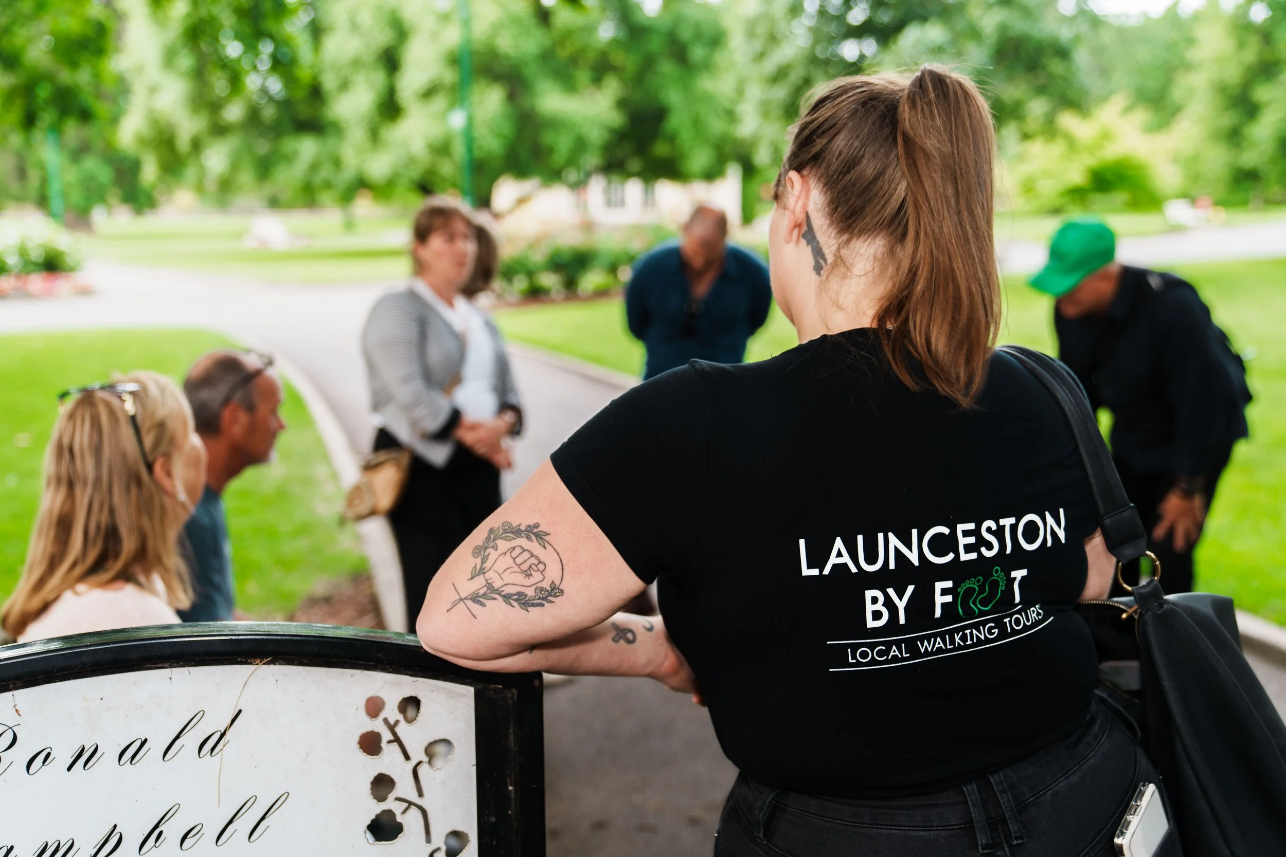 A woman with tattoos on her arm wearing a black shirt with white text, standing outdoors with people sitting and talking near a park sign with the word "board" visible. The background includes green trees and a walking path.