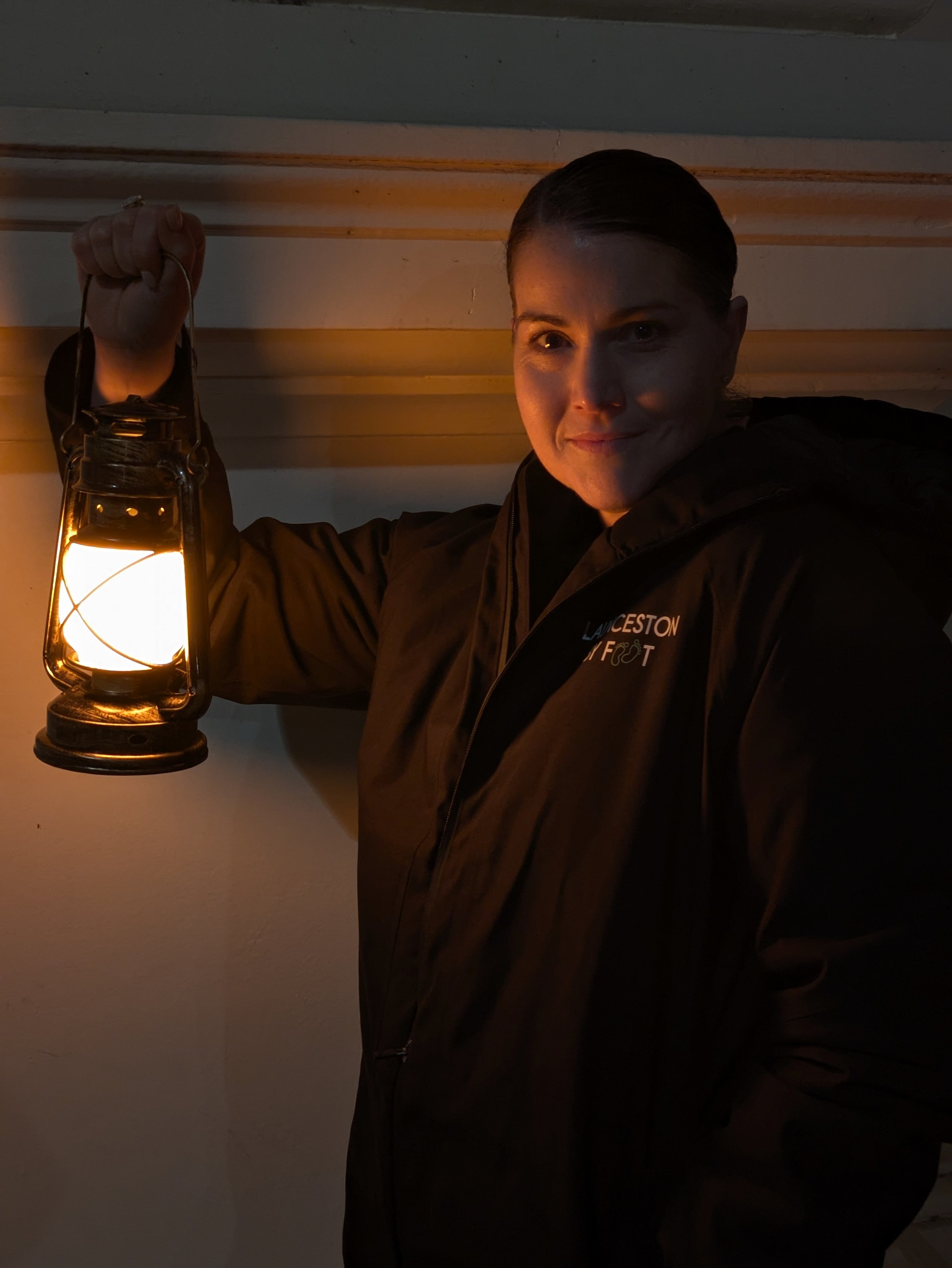 A woman holding a lit lantern and smiling at the camera, wearing a black jacket with a logo that appears to say "LANCETON FOOT." She is inside a building with painted walls and horizontal wooden trim.