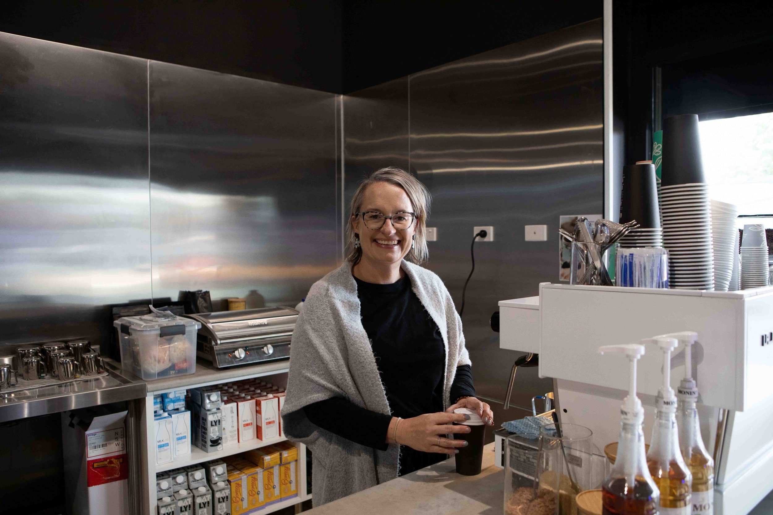 Lexi stand at the coffee machine, smiling at the camera while she steams a jug of milk