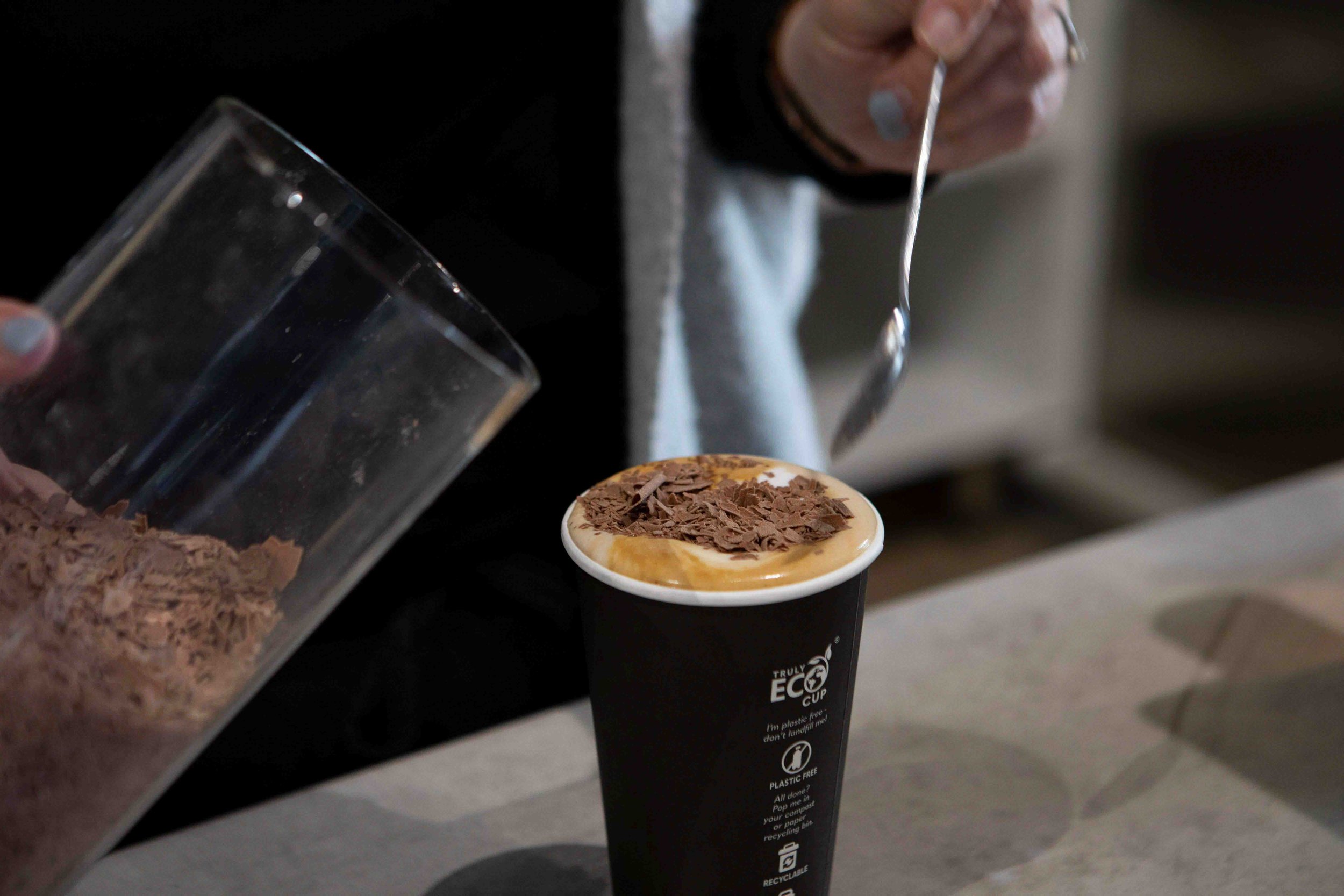 Chocolate being place on top of a cappuccino in a takeaway cup
