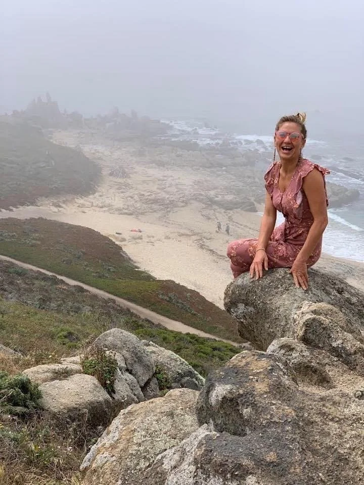 Smiling woman seated on a rock overlooking a misty beach scene far below.