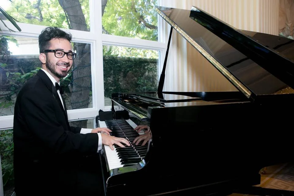 A man in a tuxedo playing a grand piano in a room with large windows showing green trees outside.