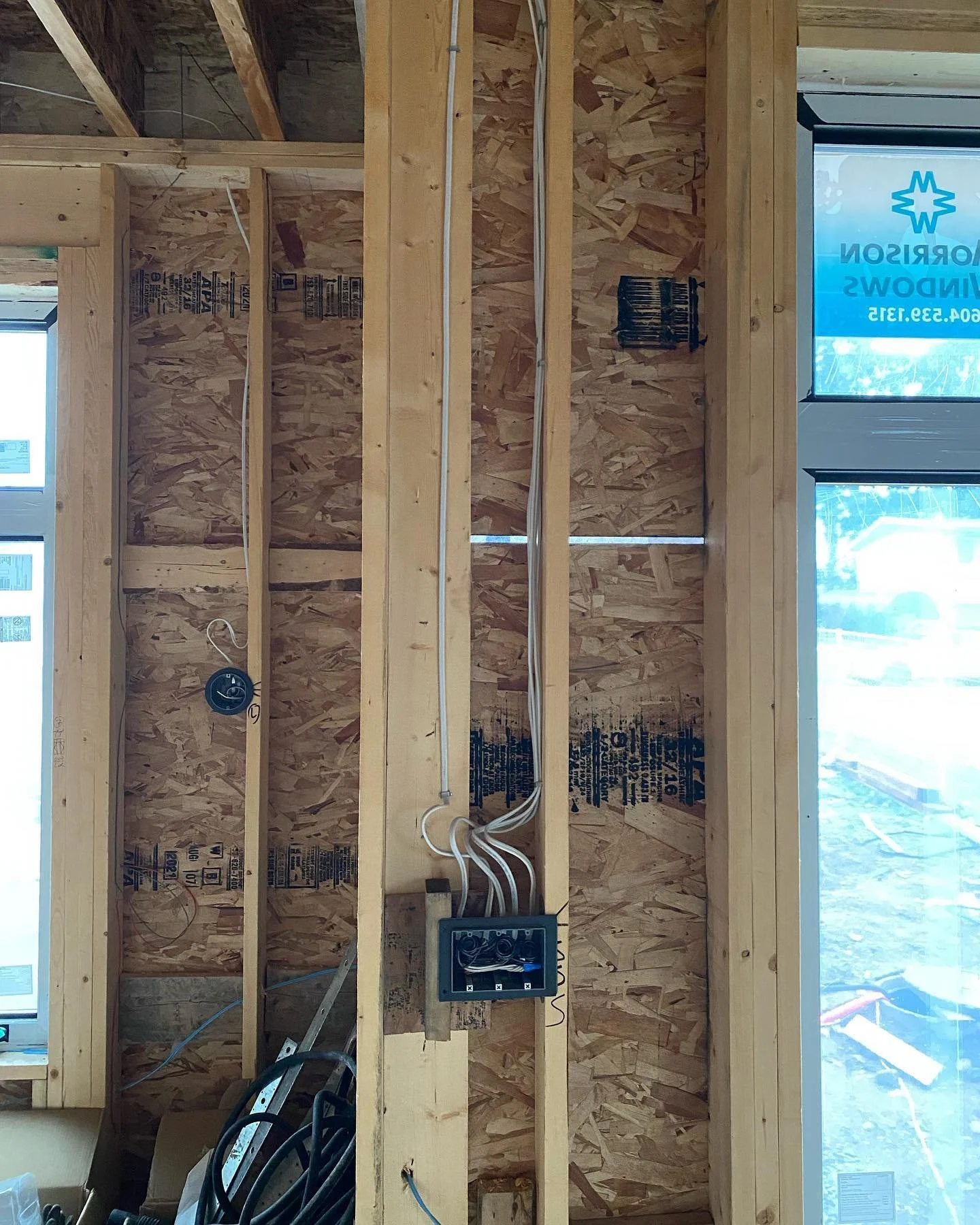 Interior of a building under construction showing exposed wood framing and OSB wall sheathing. Electrical wiring is installed with some wires hanging, and a junction box is mounted on a vertical stud. A window with a sticker is visible on the right s
