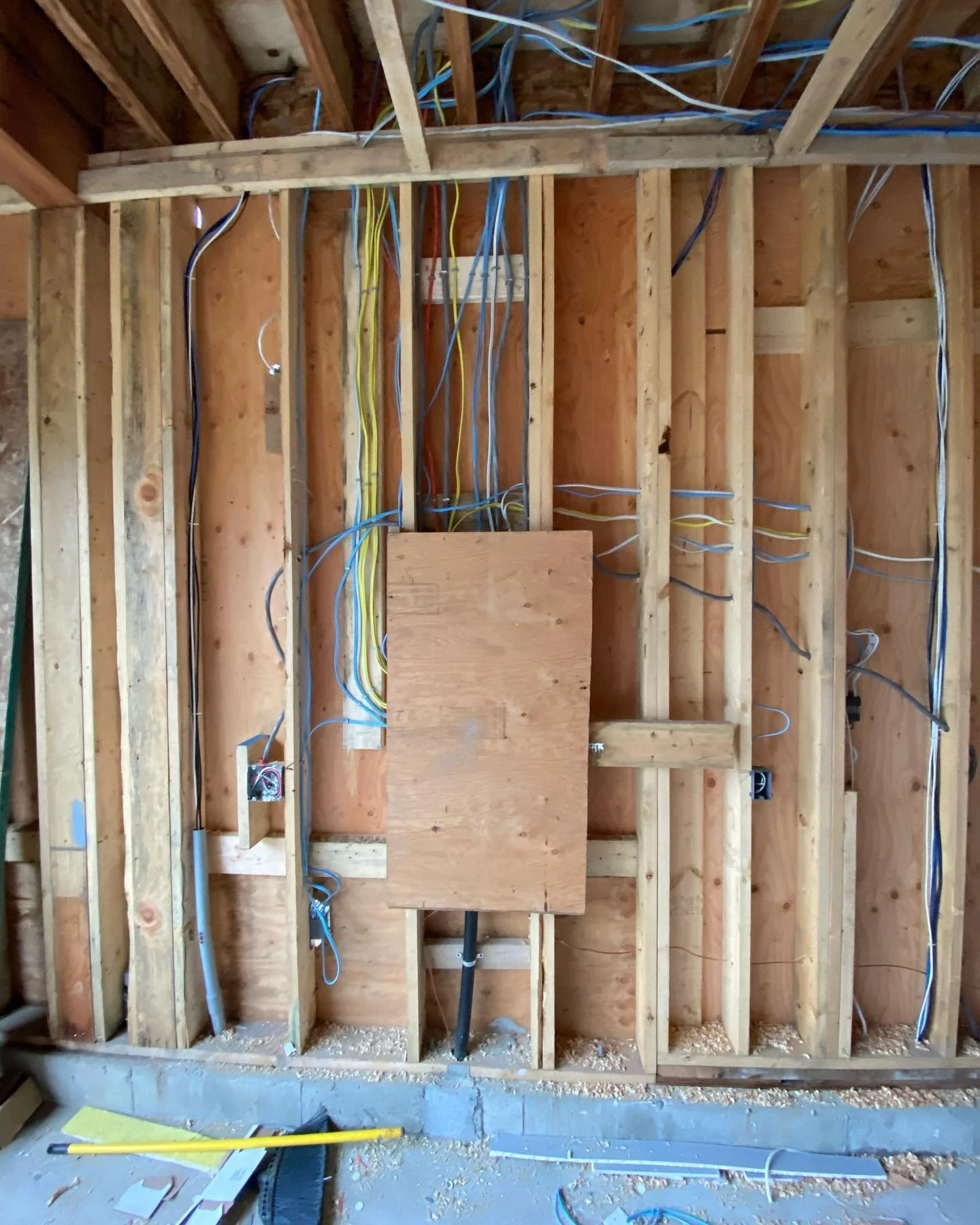 The image shows an unfinished interior wall of a building under construction, with exposed wooden studs and wiring, including yellow, blue, and white cables, and a wooden electrical box.
