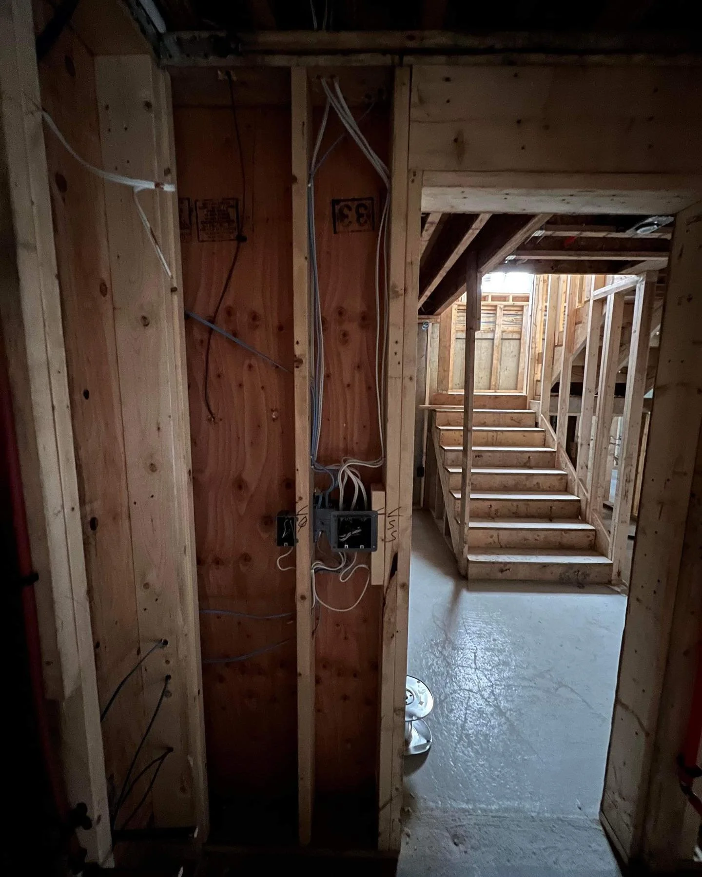 Interior view of a house under construction showing wooden framing, electrical wiring, a staircase leading to an upper floor, and a concrete floor with light coming through an opening.