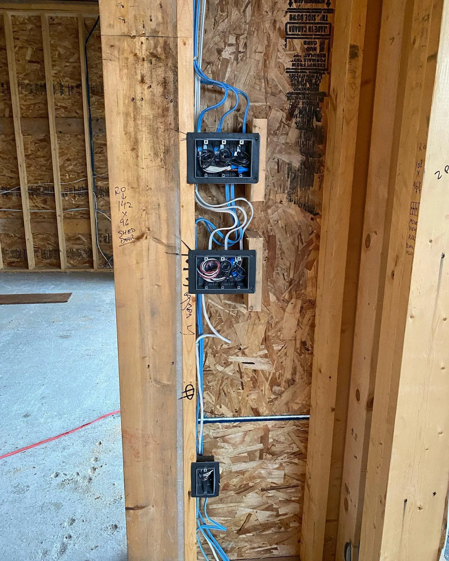 Electrical wiring setup in a construction area with two black electrical boxes mounted on a wooden wall, blue and white wires connected to the boxes, and handwritten notes on the wood.