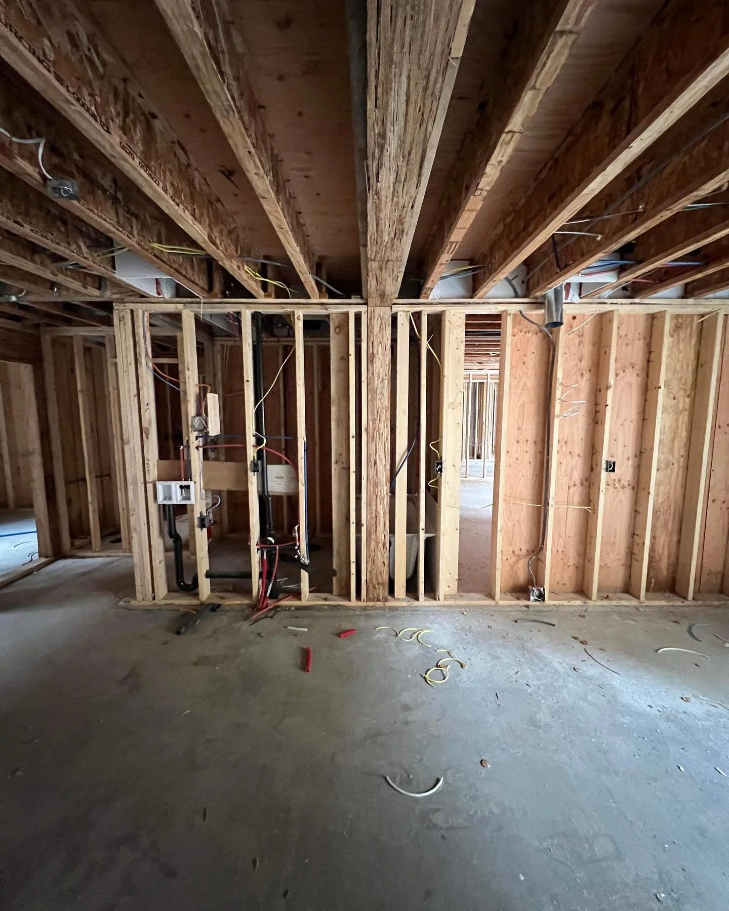 Interior of a house under construction with exposed wooden studs, wiring, and plumbing. No finished walls or ceilings are present.