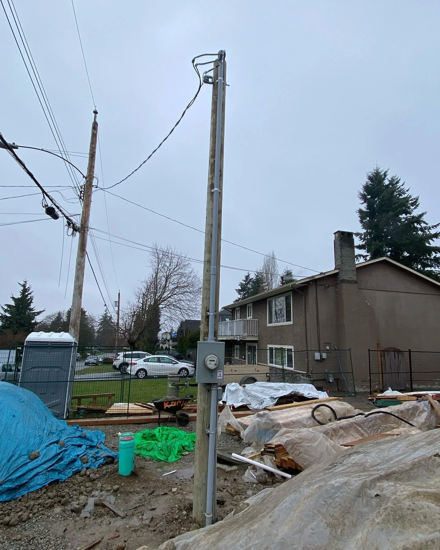 Residential backyard construction site with utility pole, construction materials, tools, and a house in the background under a cloudy sky.
