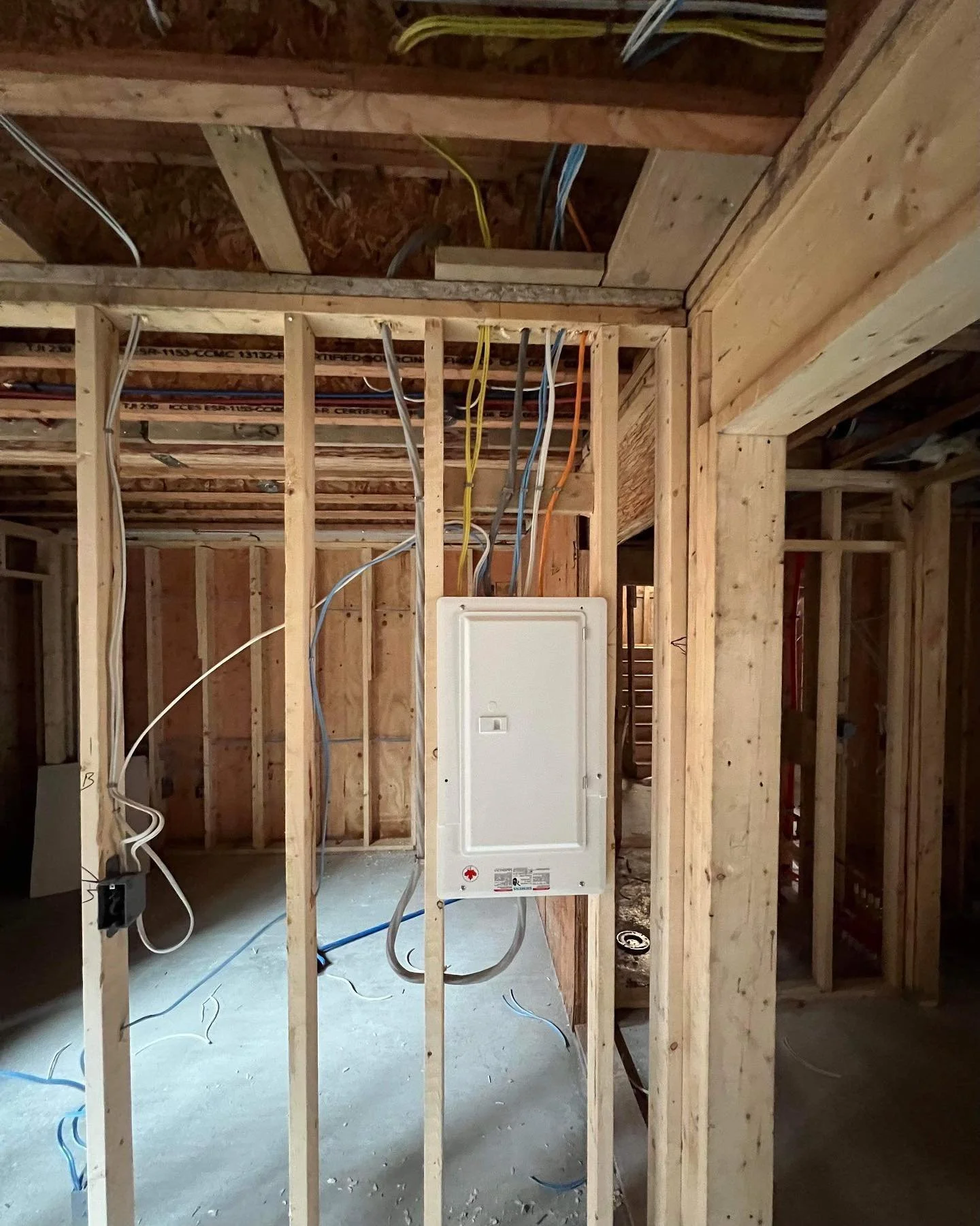 Interior of a house under construction with exposed wooden studs and electrical wiring, and a white electrical panel mounted on the wall.