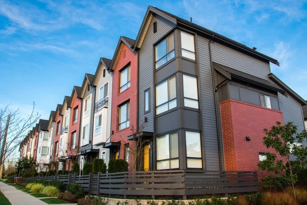 Modern multi-story apartment complex with red brick and gray siding, large windows, and small front yards with fencing and landscaping, under a blue sky.
