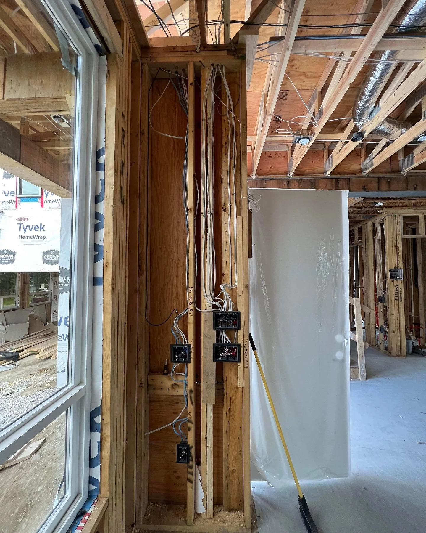 Construction site with exposed wooden studs and wiring for electrical outlets, next to a large window, with a white wall section, and exposed ceiling rafters and ductwork.