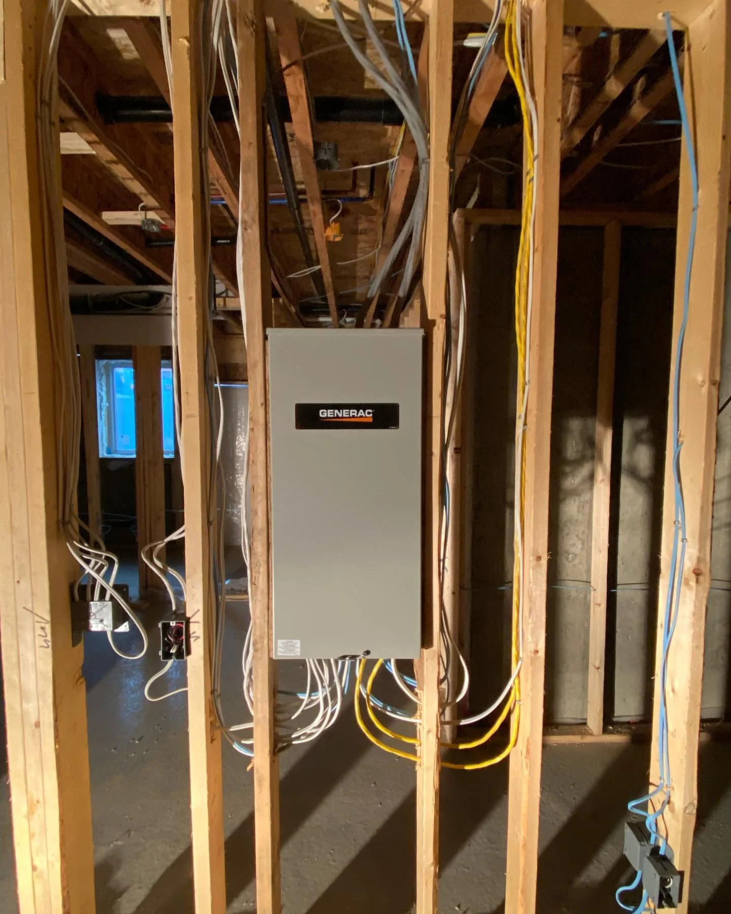 Interior view of a building under construction, showing exposed wooden framing, electrical wiring, and a gray electrical panel labeled 'Generac' mounted on the wall.