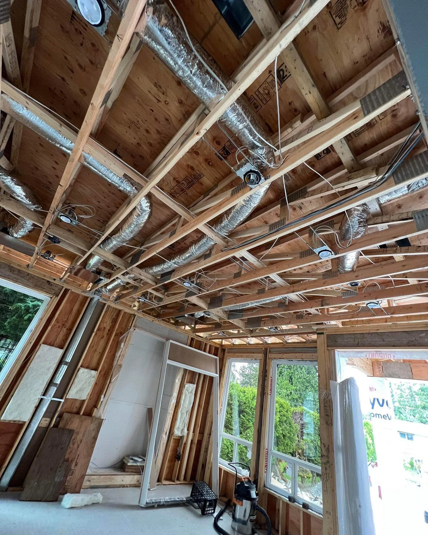 Interior of a house under construction with exposed wooden framing, air ducts, electrical wiring, and large windows.
