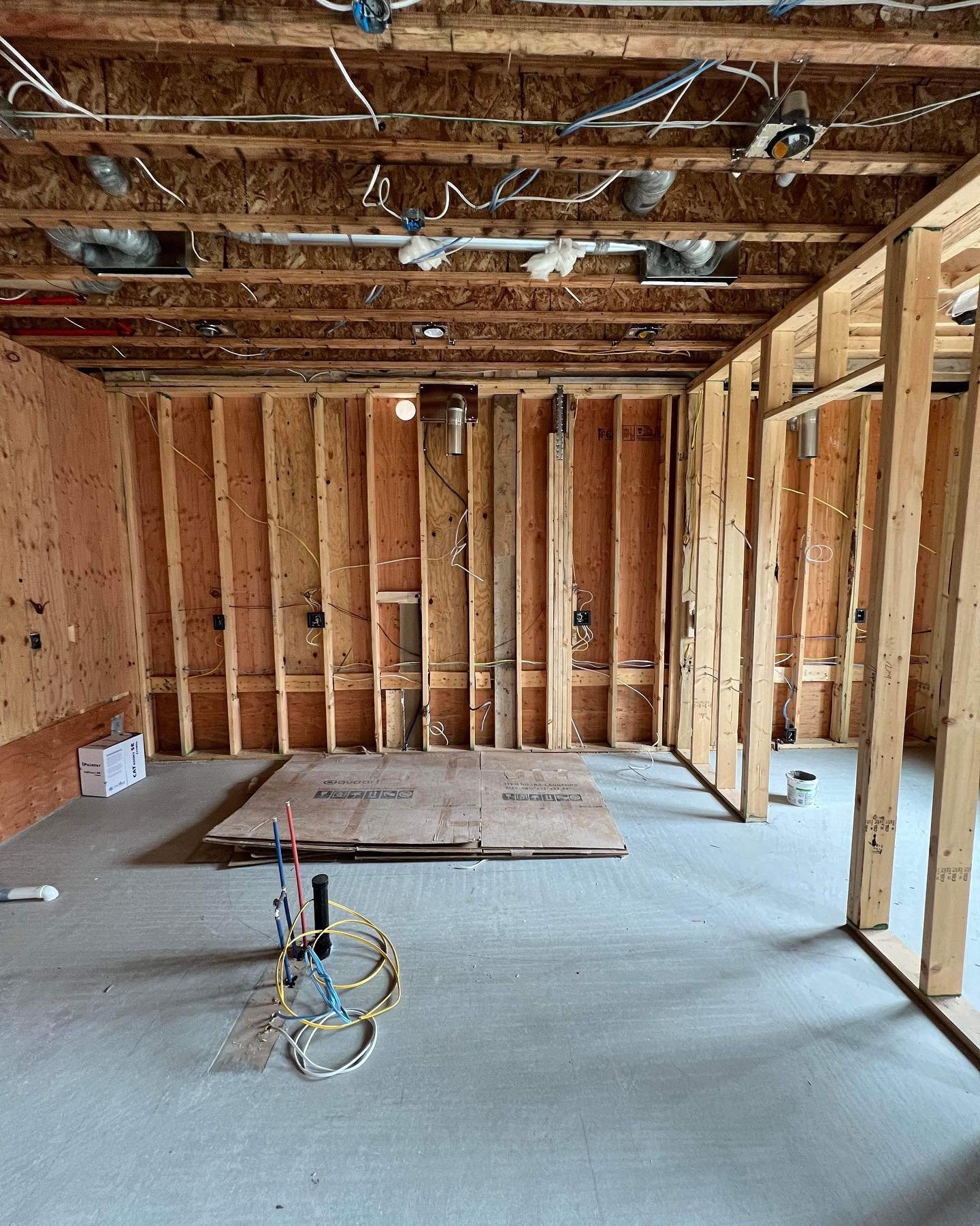 Interior of a room under construction with exposed wooden framing, electrical wiring, and HVAC ductwork. The floor has some cardboard and construction materials.