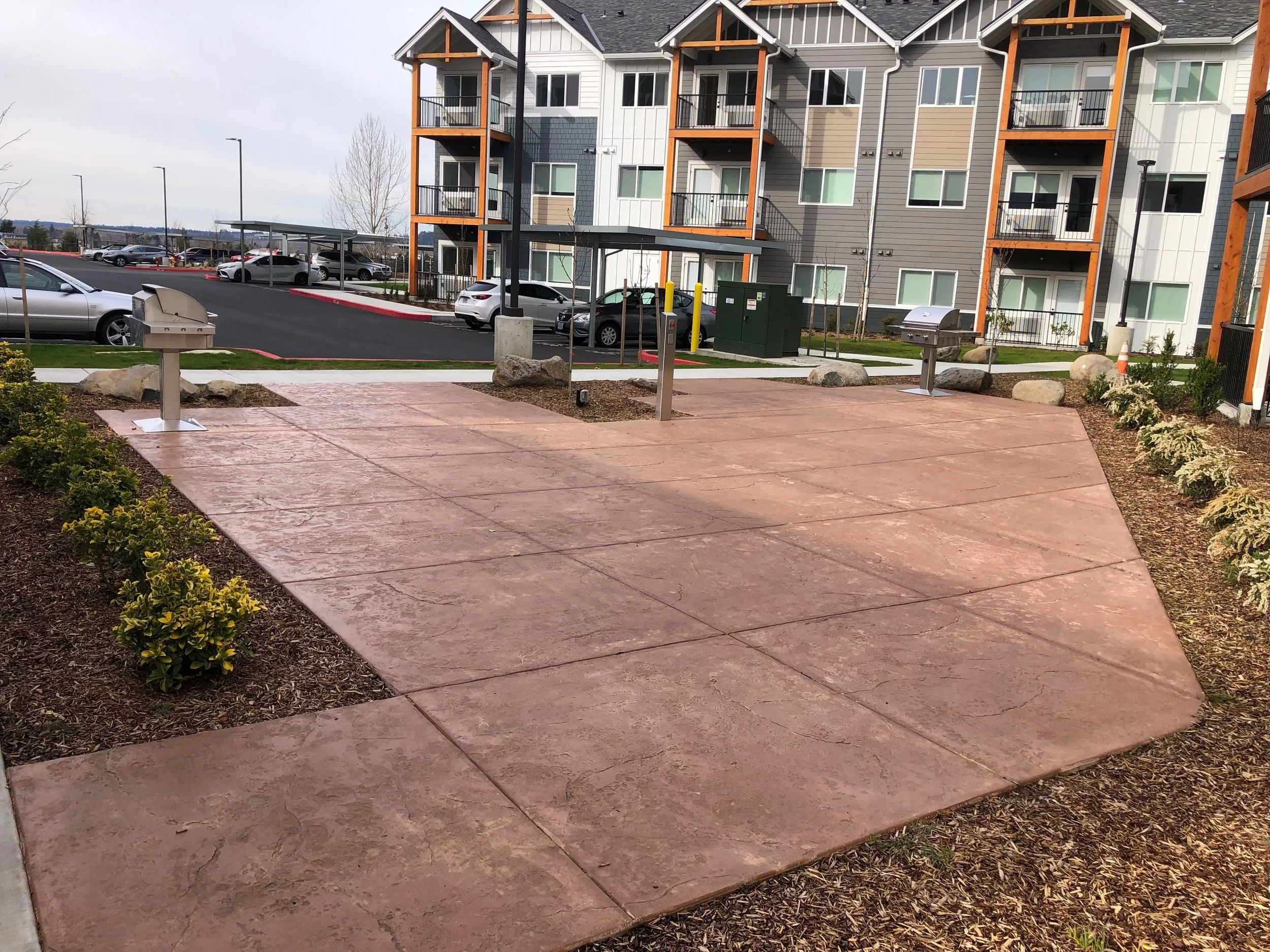 A stamped concrete walkway in front of a multi-story apartment building with balconies. There are cars parked along the curb, small trees, and a landscaped area with bushes and rocks. The sky is cloudy.