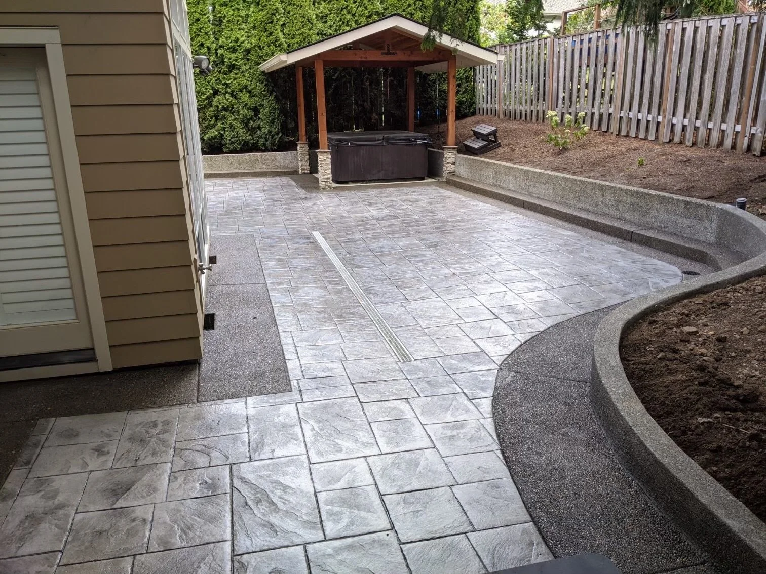A newly paved backyard patio with a hot tub under a wooden gazebo, a wooden fence, a garden bed, and a drain system in the concrete surface.