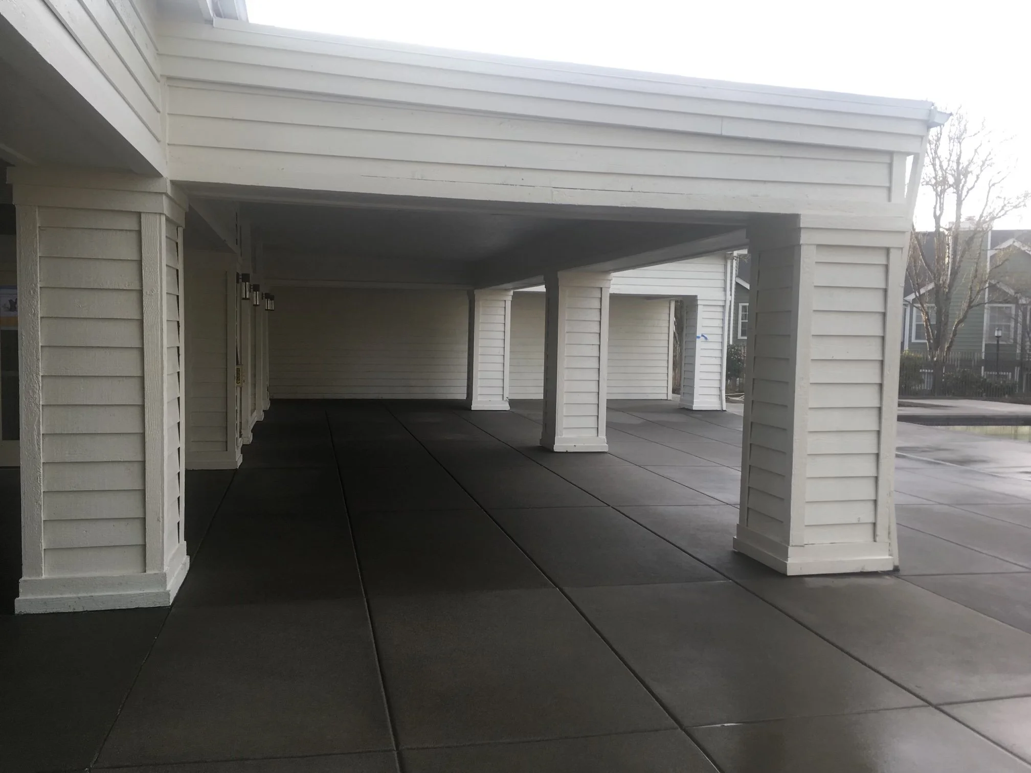 View of a covered porch area with white wooden siding, ceiling, and columns, with an outdoor scene of leafless trees and neighboring houses visible in the background.