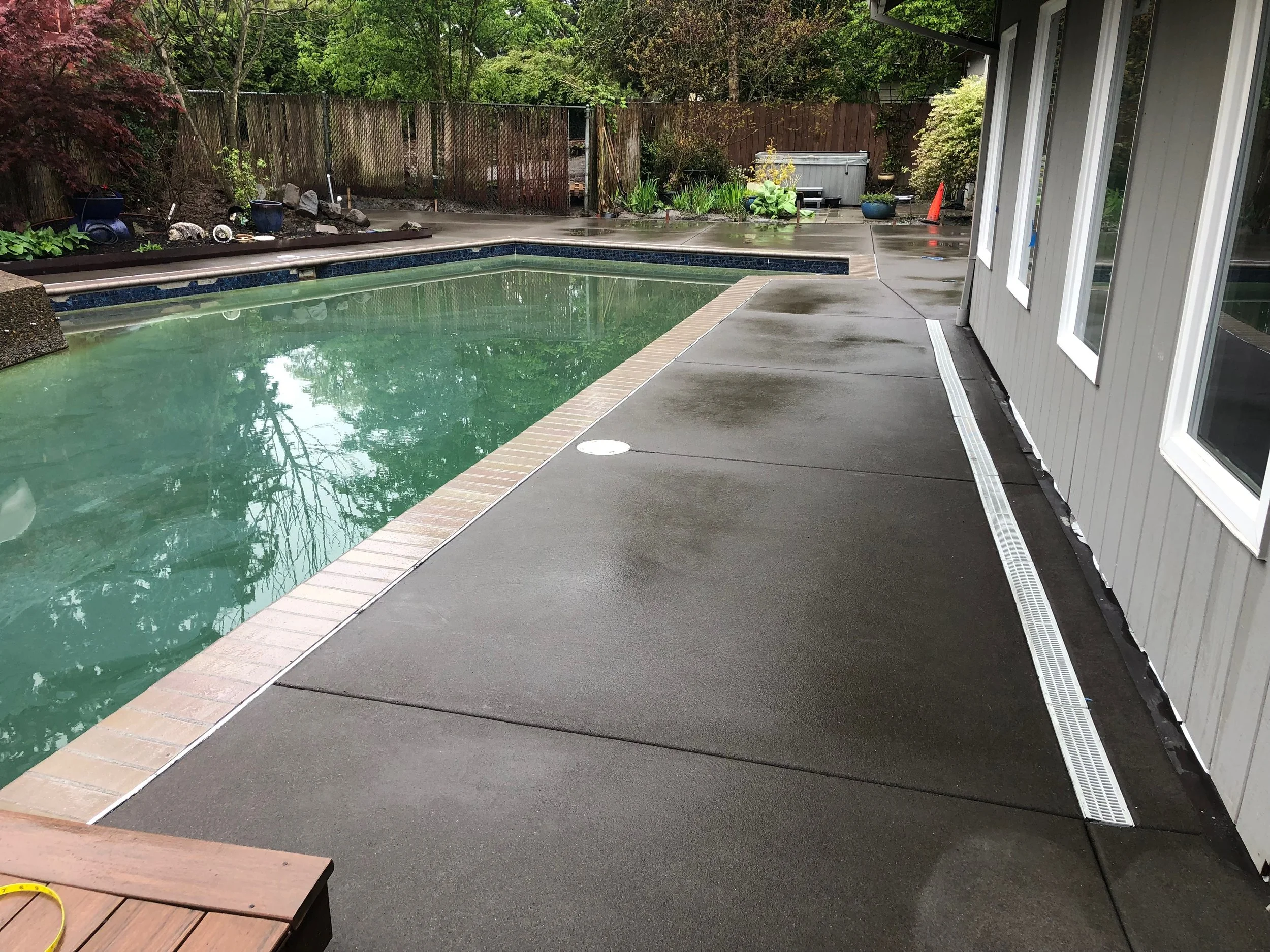 Backyard with a swimming pool, wet concrete patio, plants, and trees, next to a house with a gray exterior and large windows, wet from rain.