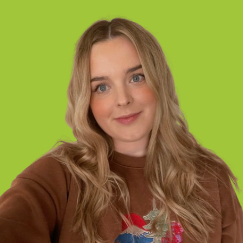 A young woman with long blonde hair, blue eyes, and a light complexion, wearing a brown sweatshirt with a colorful graphic, posing against a bright green background.