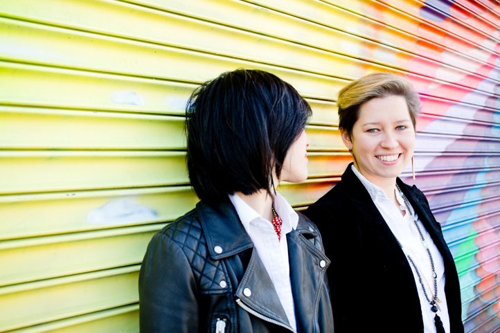 Alice &amp; Catie look at each other and the camera smiling with a colorful mural in the background in Bushwick, Brooklyn