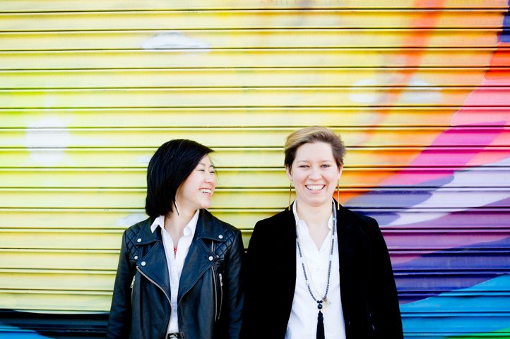 Alice &amp; Catie look at each other and the camera smiling with a colorful mural in the background in Bushwick, Brooklyn