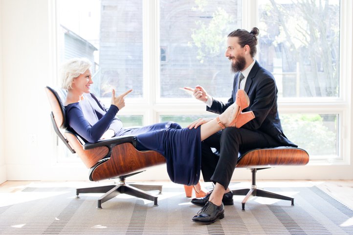 Randy &amp; Kelsey Taylor sit in office chairs point at each other with finger guns in their home in Hudson, NY