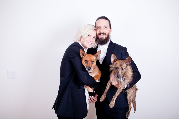 Randy &amp; Kelsey Taylor stand smiling, holding their dogs while wearing suits in their home in Hudson, NY