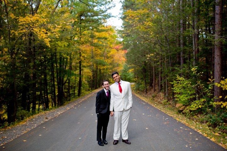 Arthur &amp; Scott hold hands and smile at the camera on a wooded street in Hudson Valley, NY during the fall