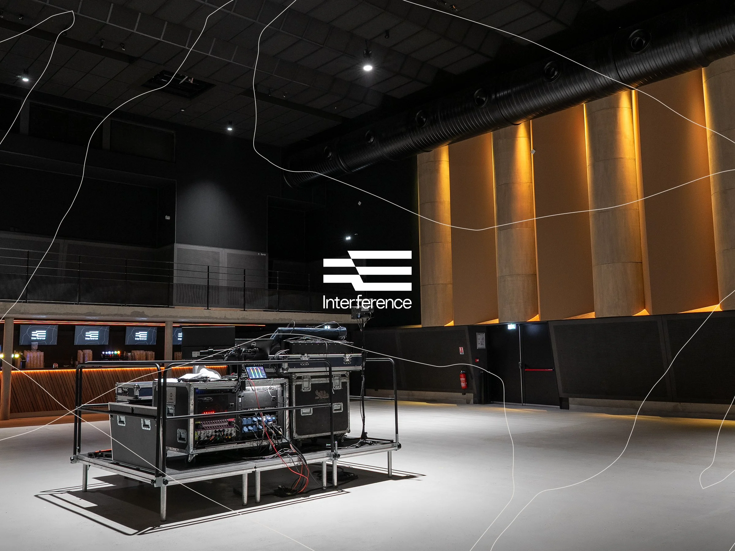 Une scène intérieure d'un auditorium ou d'une salle de spectacle avec un plafond noir, des colonnes en béton éclairées par une lumière chaude, un système de sonorisation suspendu, et un équipement technique de performance ou de sonorisation placé sur