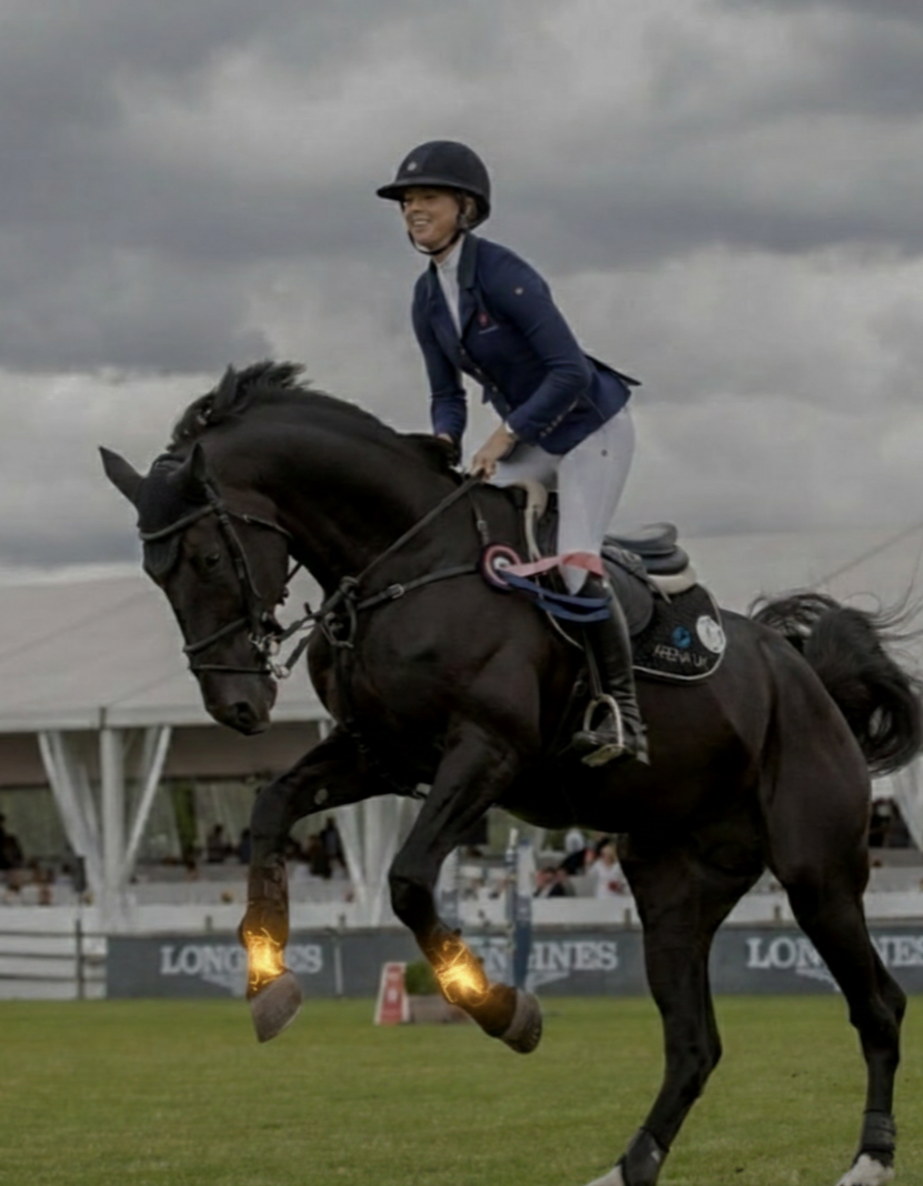 Eine Reiterin in Reitkleidung auf einem springenden schwarzen Pferd während eines Wettbewerbs bei bewölktem Himmel.