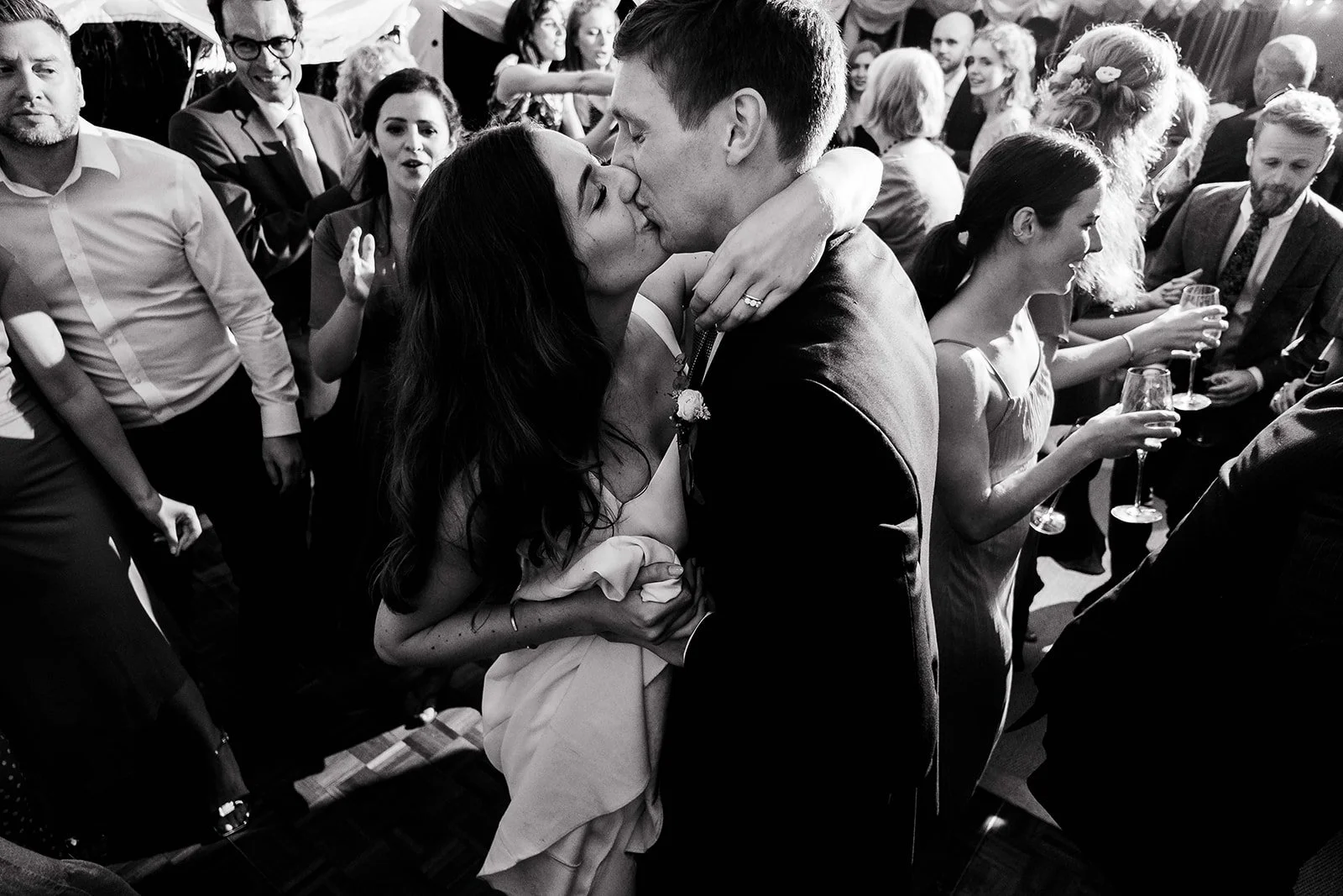 A couple sharing a kiss at a wedding reception with guests surrounding them, holding glasses of wine.