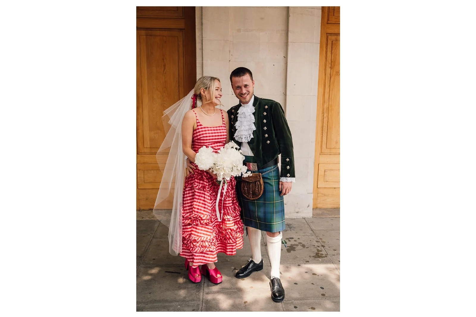 A man and woman in traditional Scottish attire standing together outside Hackney town hall, smiling. The woman is wearing a red and white checkered dress, holding a bouquet of white flowers, and has a veil.