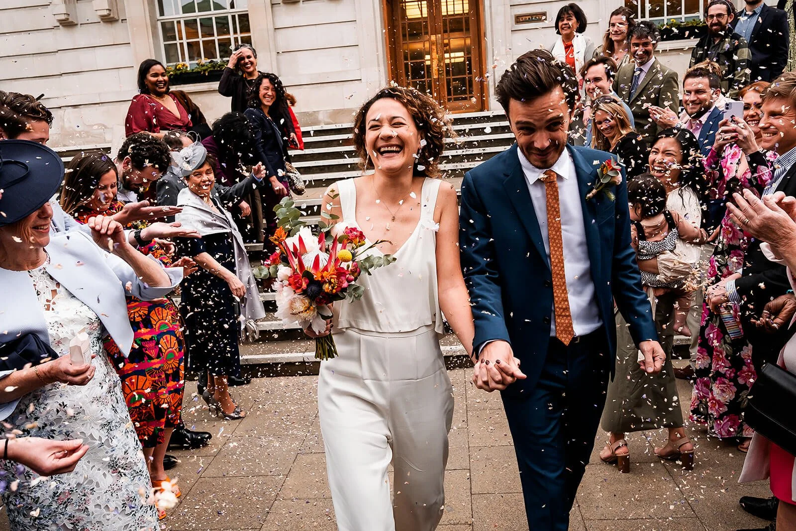 A newlywed couple hand-in-hand walking down the steps outside hackney town hall, smiling and celebrating with friends and family throwing confetti.