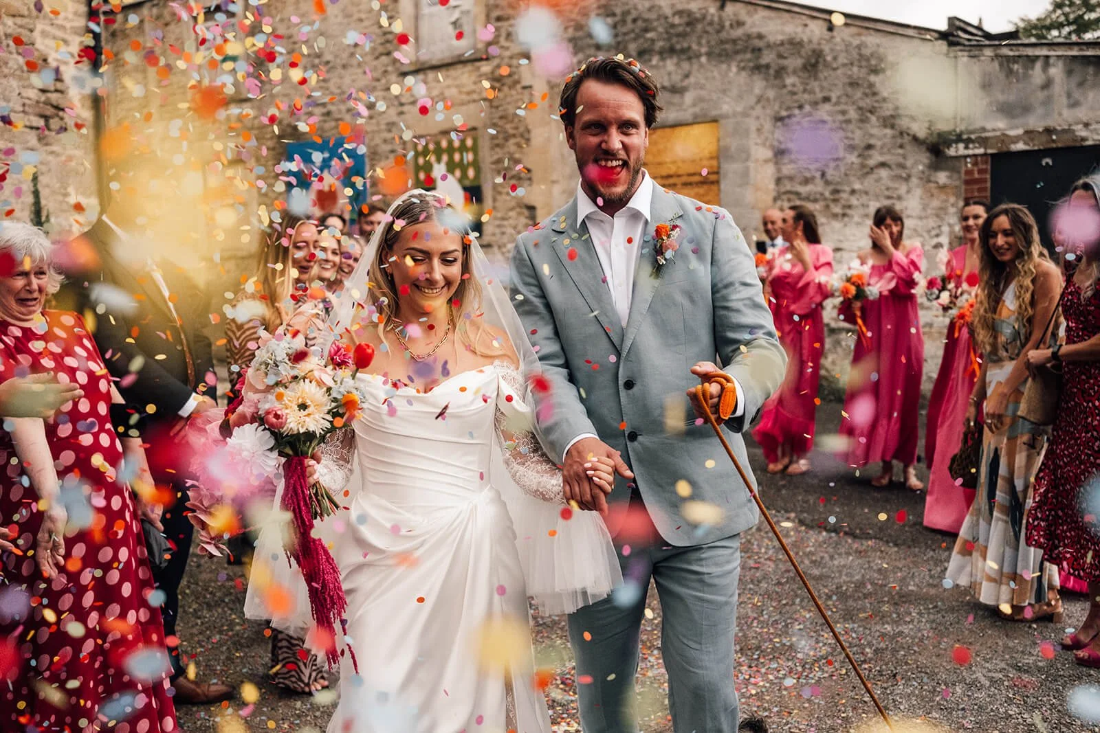 A newlywed couple walking hand-in-hand through a celebration with colorful confetti. The bride is wearing a white wedding dress and holding a bouquet, and the groom is in a light grey suit with a boutonniere. Guests in bright dresses are cheering and clapping around them in an outdoor setting with a rustic wall background.