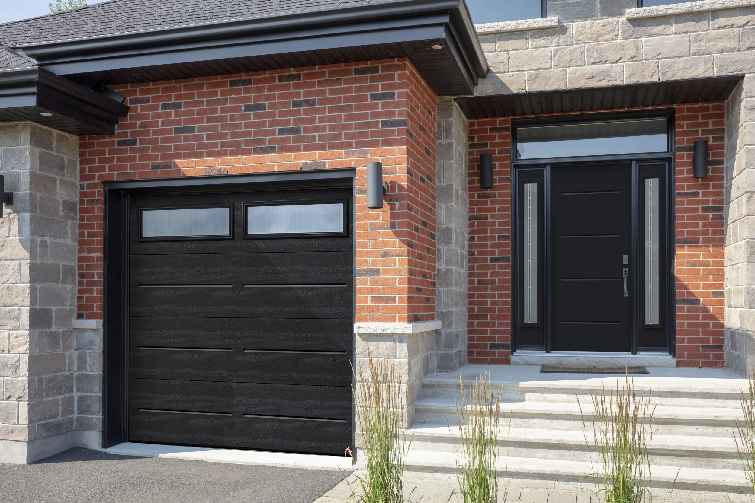 Modern house entrance with a black front door and a garage door, brick and stone exterior, steps leading to the door, and small landscaping plants.