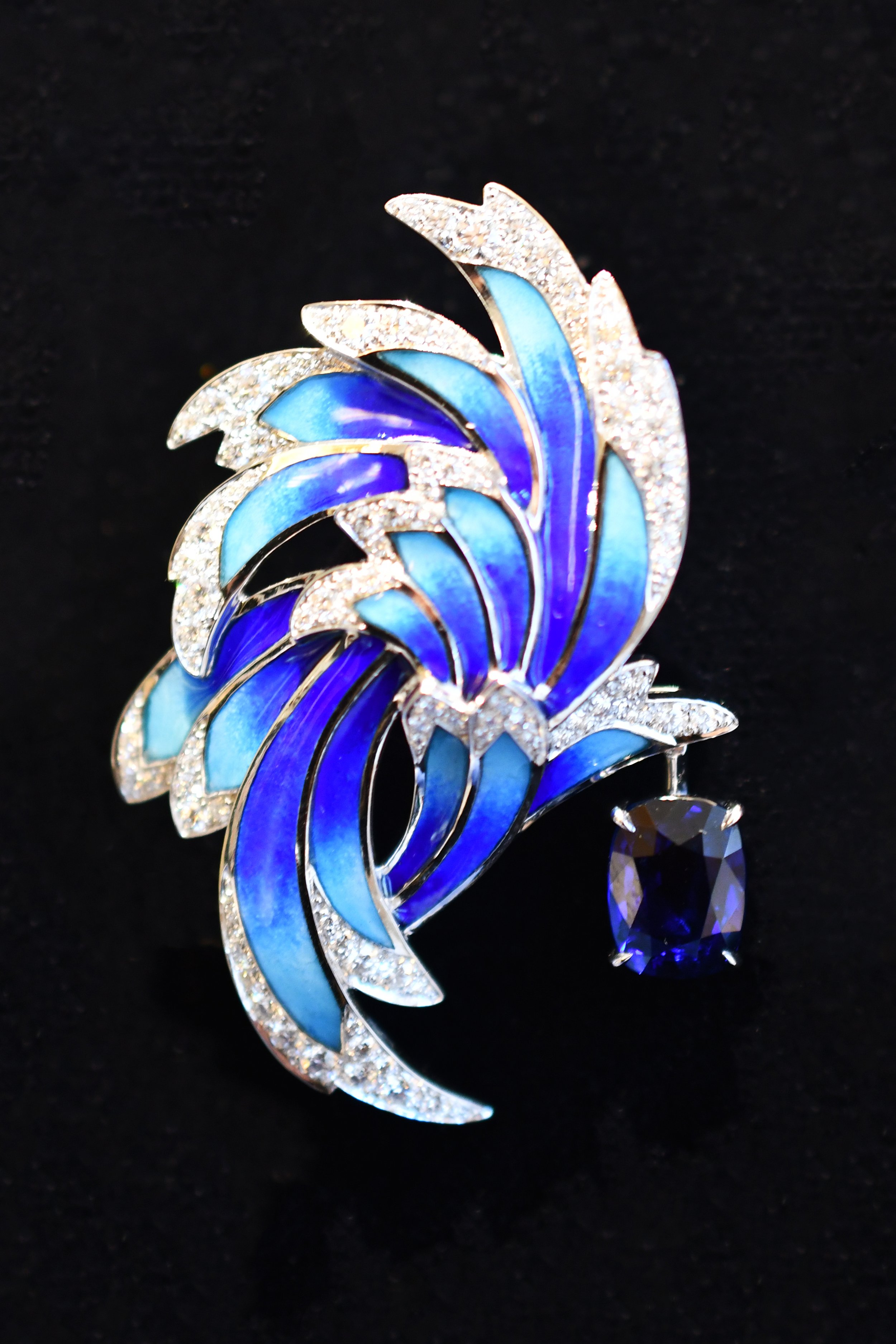 A decorative brooch with a floral design, featuring blue and light blue enamel petals and a dangling dark purple gemstone, set against a black background. Chaumet’s Envol Collection