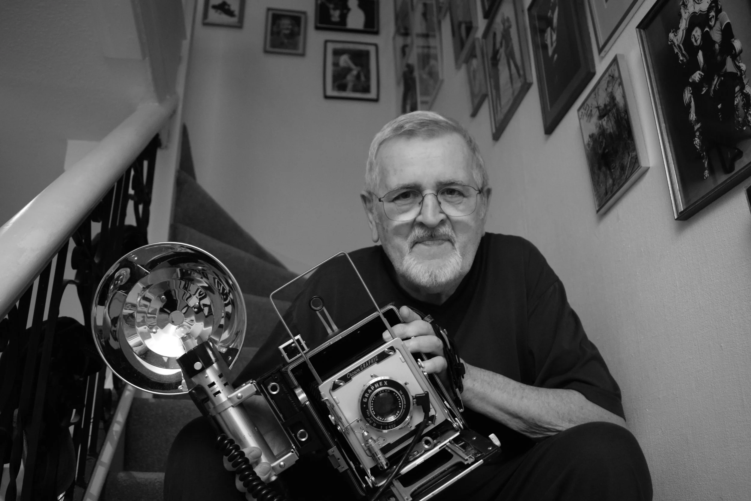 A black and white photo of an older man with glasses and a beard, sitting on a staircase holding a vintage large-format camera.