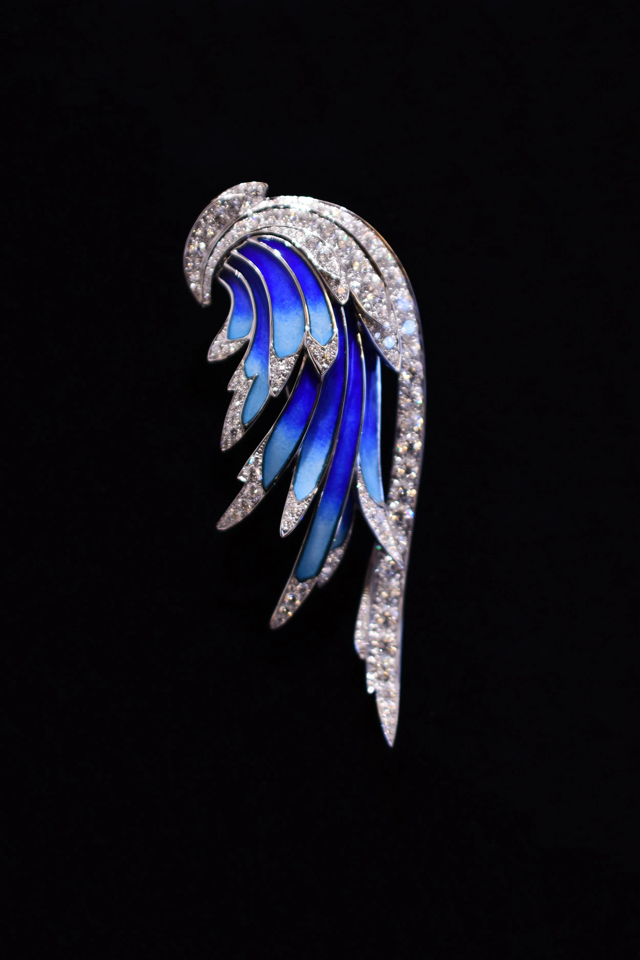 A silver and blue feather-shaped jewelry piece with diamonds on a black background. Chaumet’s Envol Collection