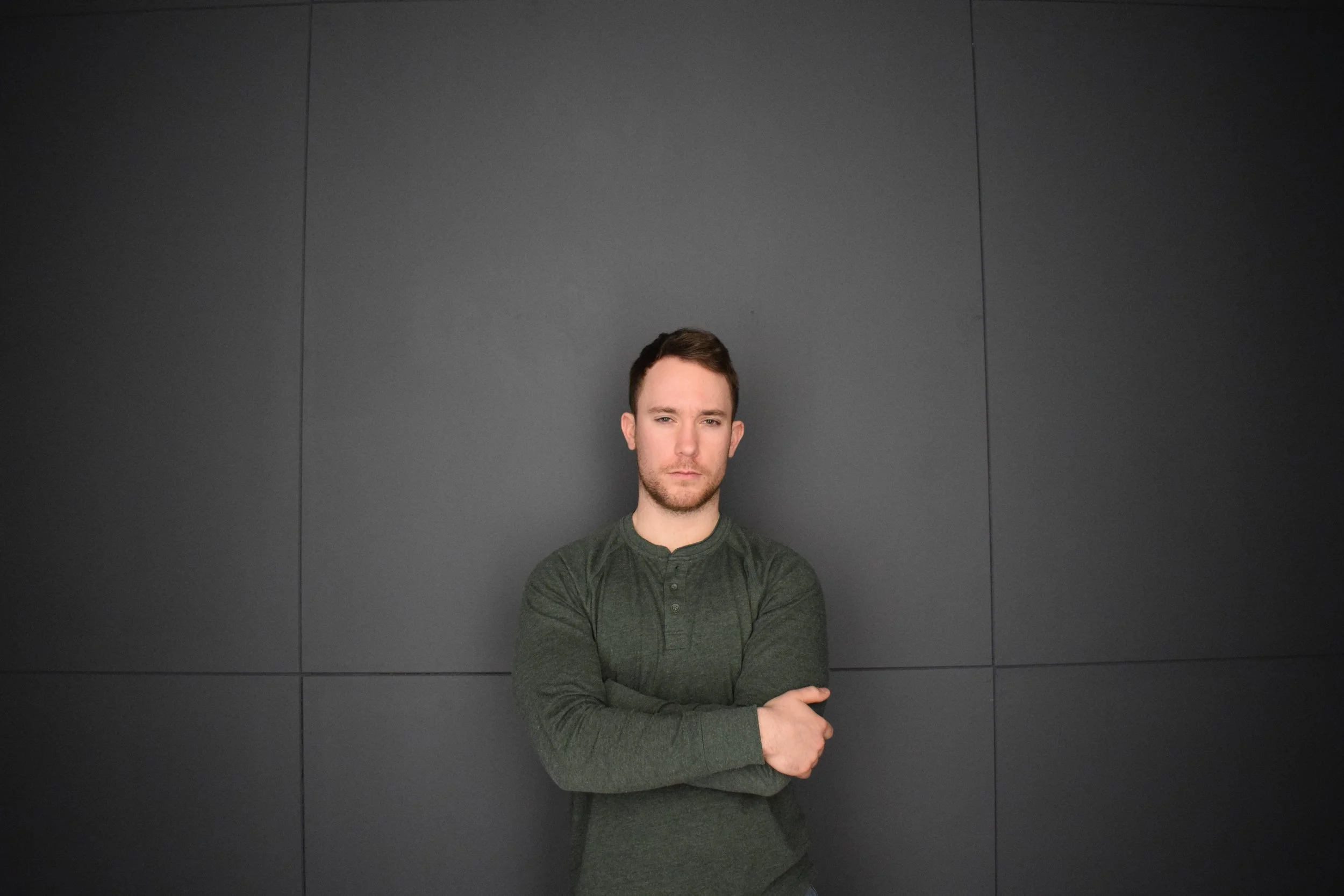 A man with brown hair and a beard standing against a dark gray wall, crossing his arms, wearing a long-sleeve green shirt.
