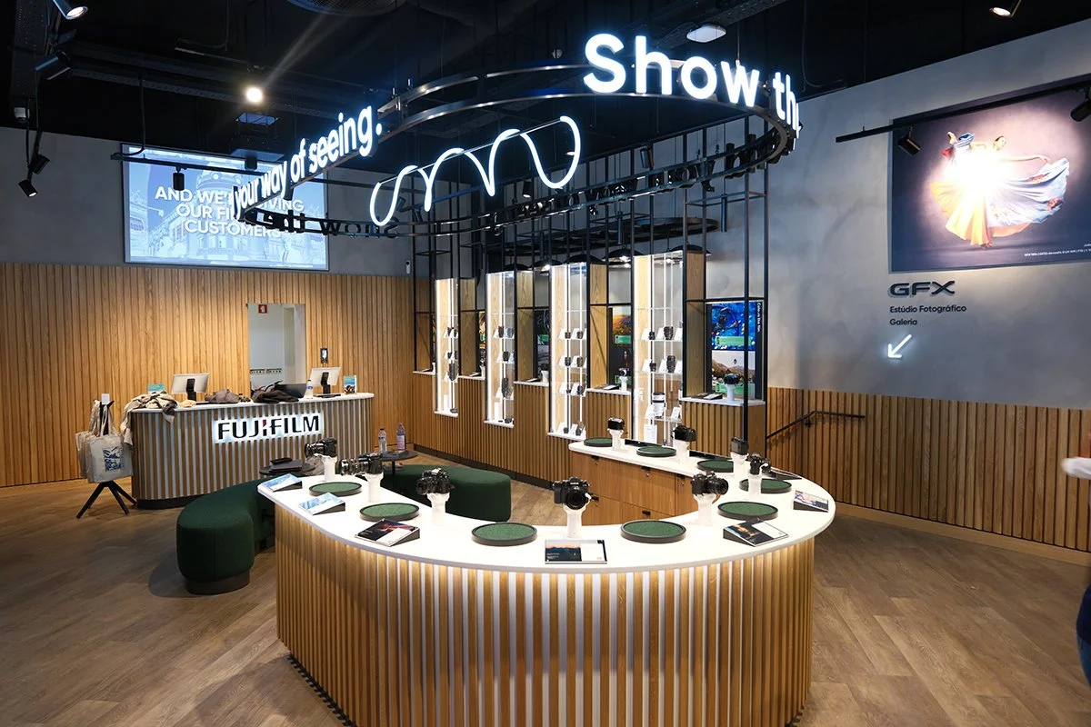 Interior of a camera store with a circular display table showcasing cameras and accessories, a counter labeled FUJIFILM, wooden wall panels, neon sign overhead, and a large camera advertisement on the wall.