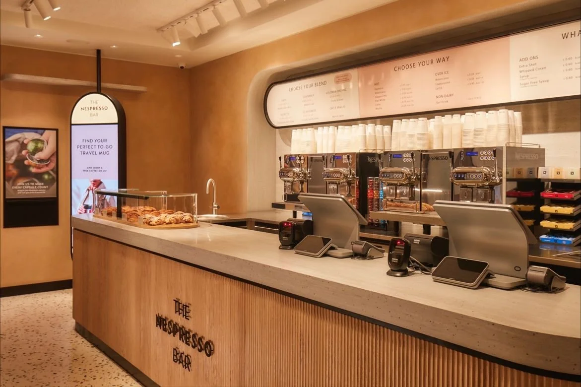 A coffee shop counter with espresso machines and cups, a sign for The Nespresso Bar, and a display case with pastries.
