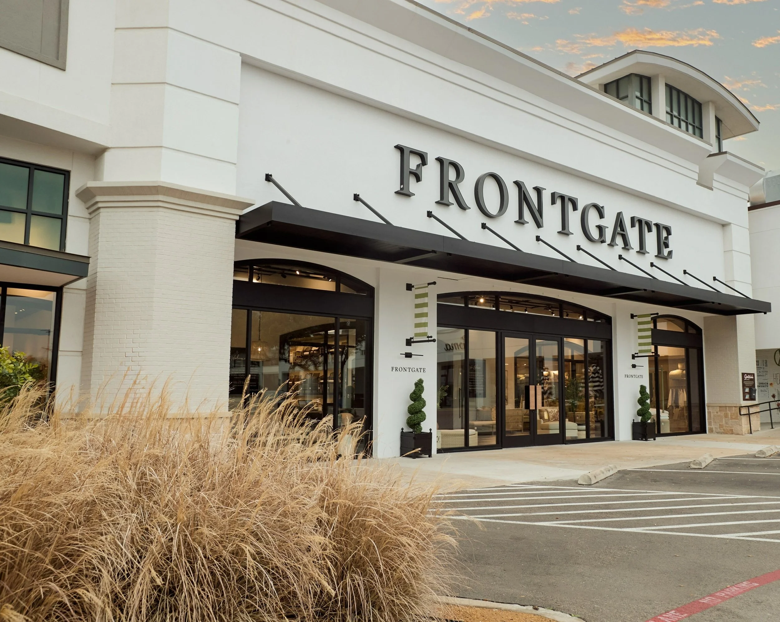 Exterior view of a retail store named Frontgate with large glass doors, white facade, and black signage, surrounded by dry ornamental grass and parking spaces.