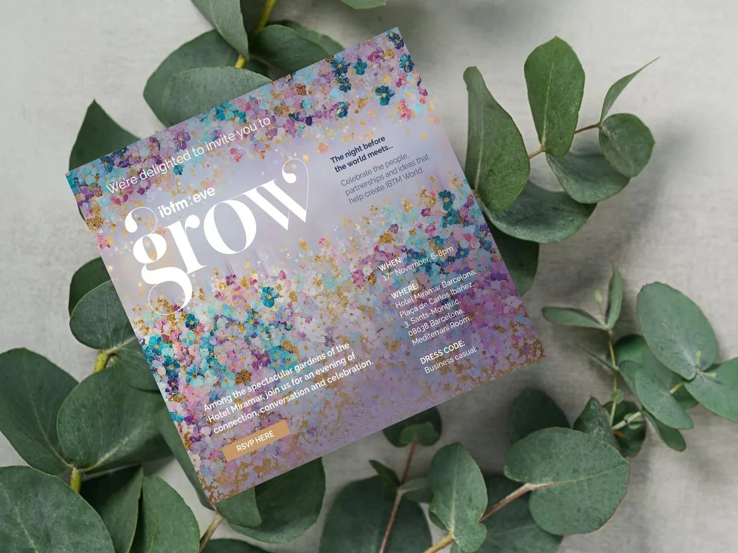 Event invitation card with colorful floral design and white text, placed on green eucalyptus leaves.