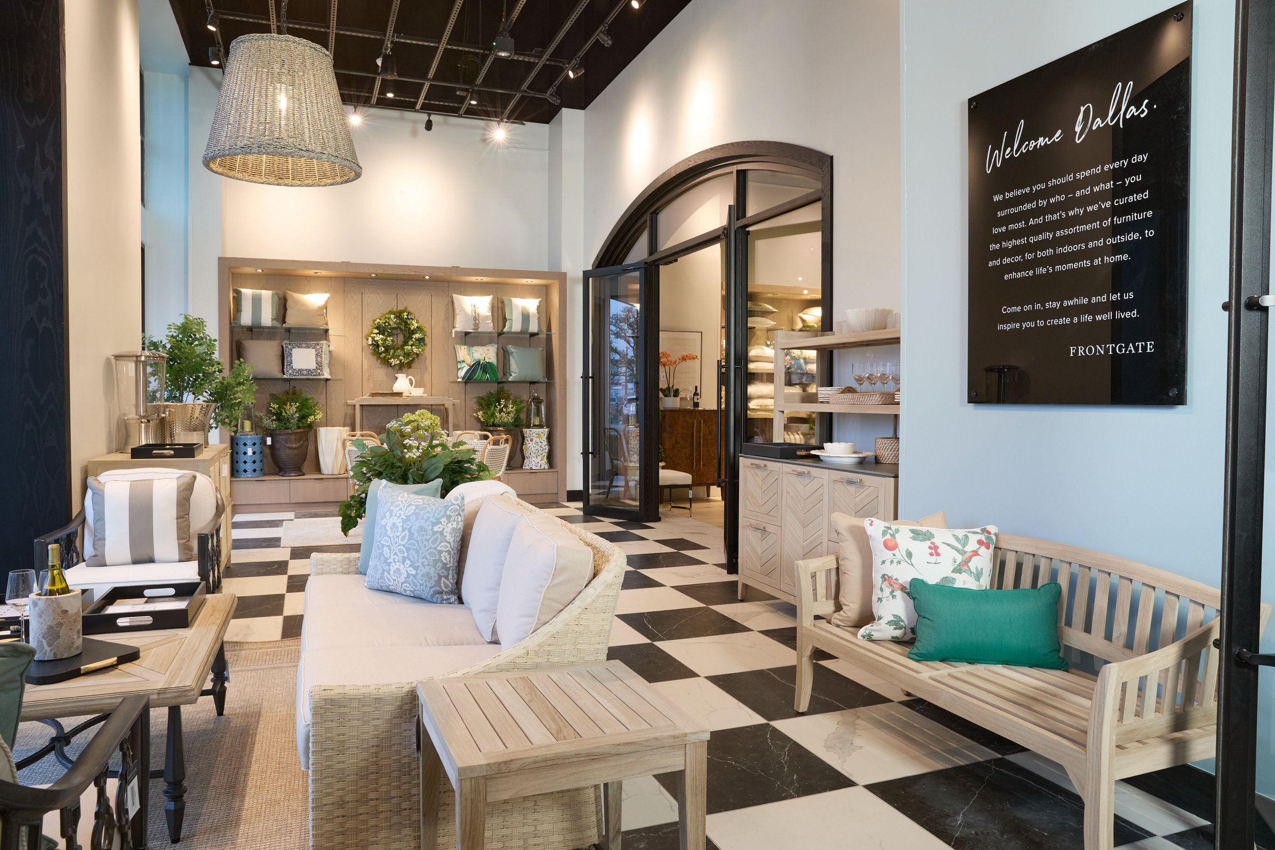 Interior of a furniture store with a seating area featuring white and light wood furniture, decorative pillows, and plants. There is black and white checkered flooring, a large woven lamp hanging from the ceiling, and a black sign on the wall that reads "Welcome Dallas."
