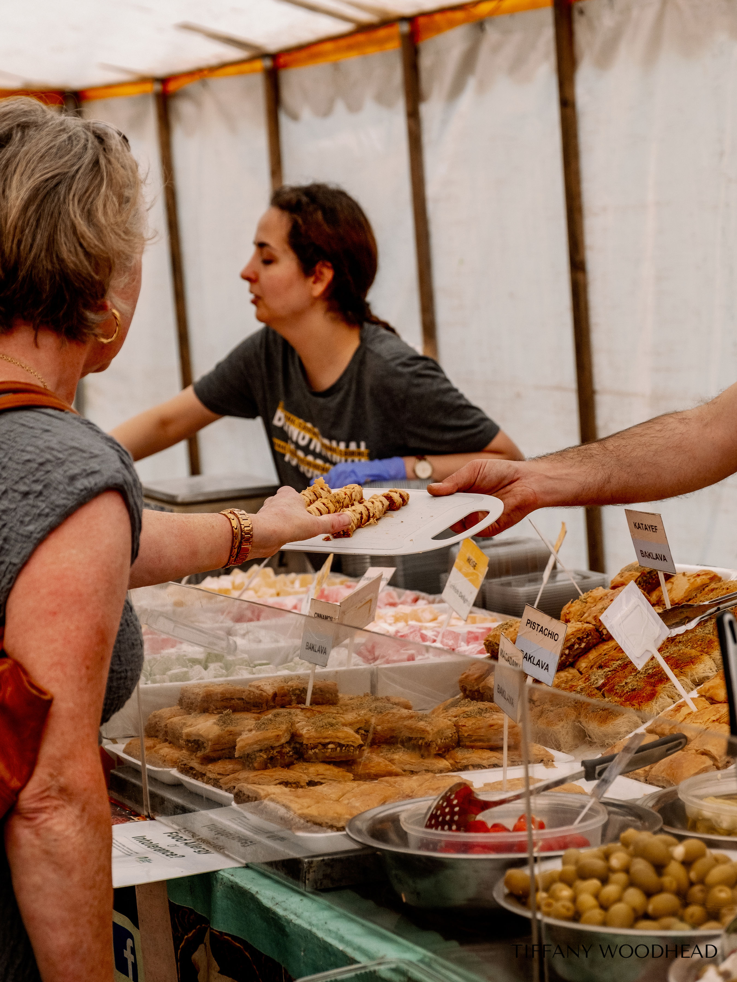 A woman at a food stall selecting baked goods from trays with labels, with other customers and food items on display in the background.