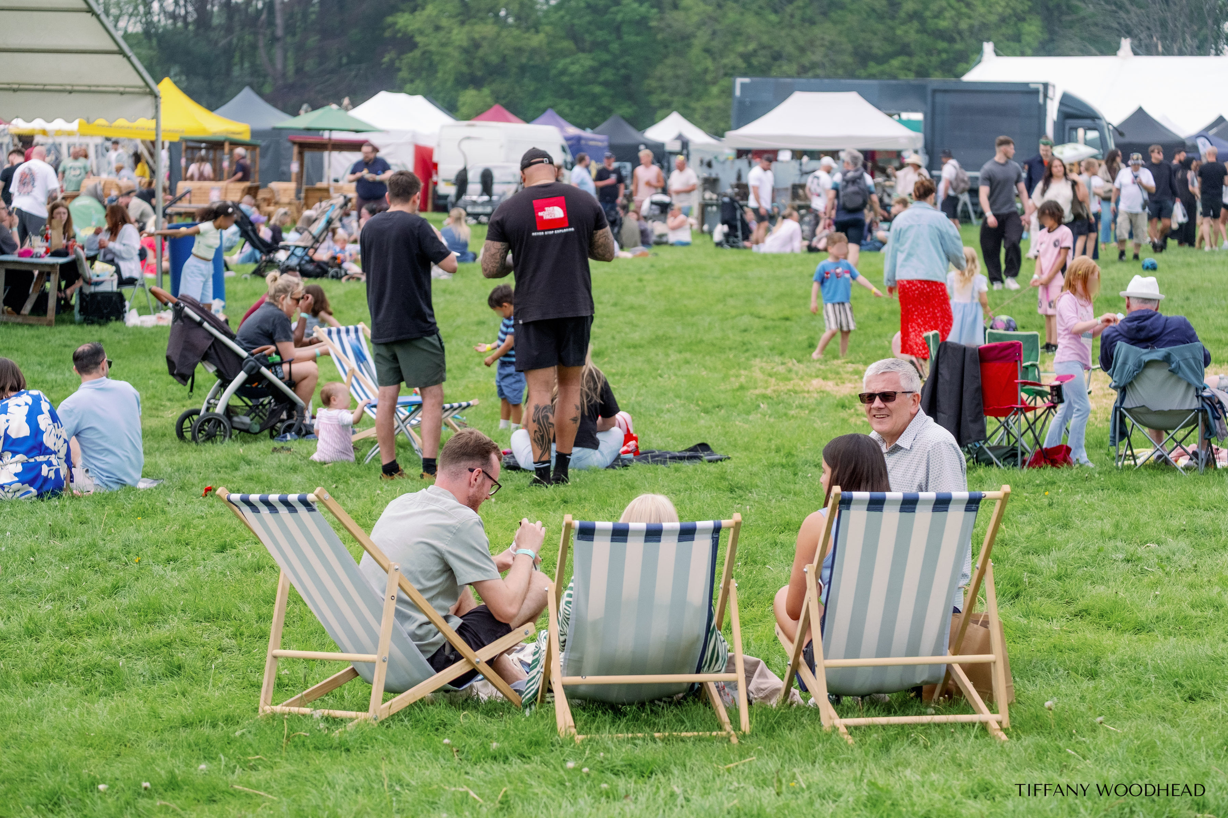 People gathering outdoors on a grassy field at a festival or fair, with tents and food trucks in the background. Some are sitting in chairs, while others stand, walk, or look at their phones. Children are playing and walking around, and adults are ch