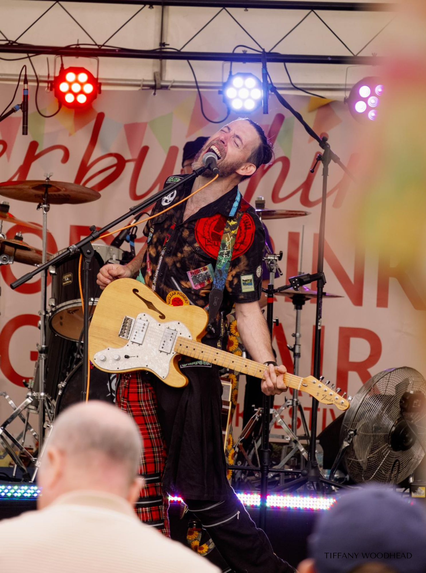 A male musician passionately singing into a microphone while playing an electric guitar on stage. Behind him, there is a drum set and colorful stage lights. The background has a banner with partial red text and triangular decorations. Audience member
