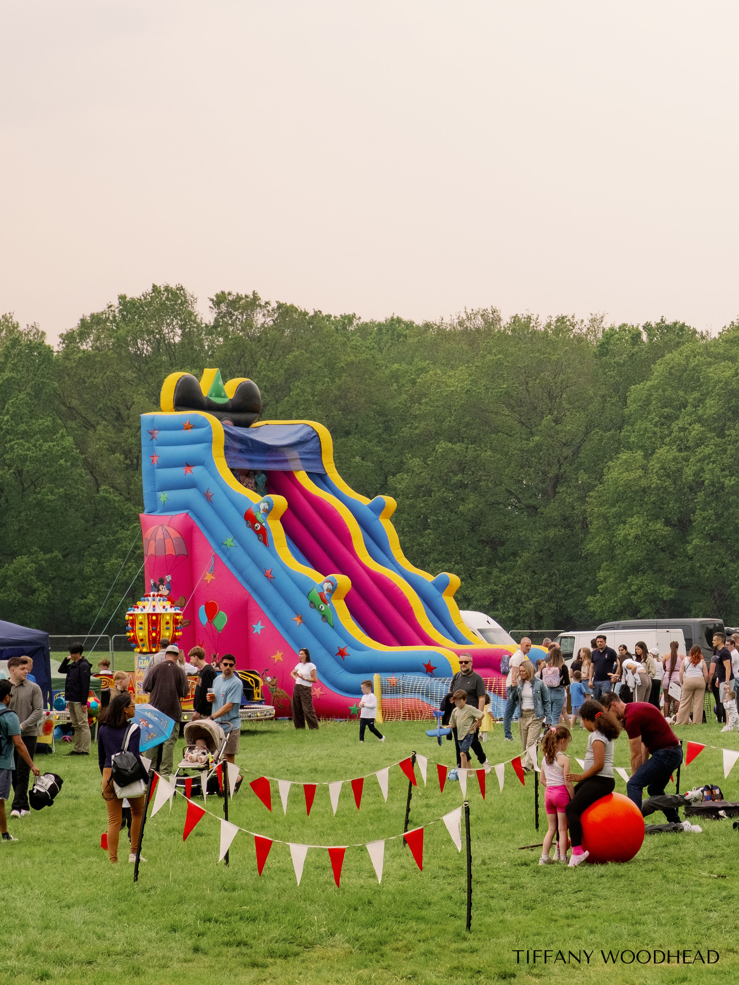 People at a fairground with a large inflatable slide featuring a cartoon character in the top section, and various children and adults walking around the green field area, with striped red and white banners in the foreground and trees in the backgrou