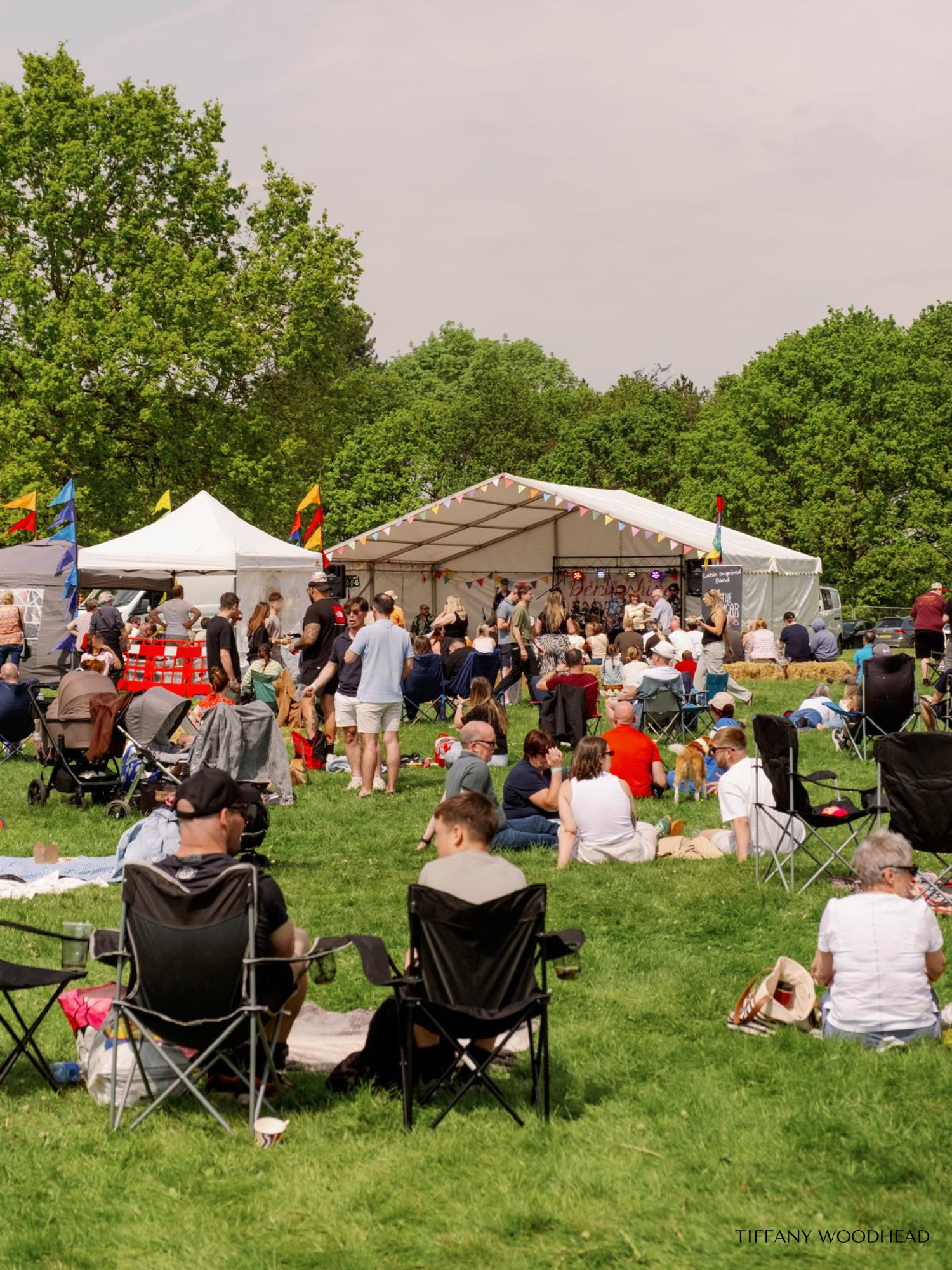Outdoor concert with a crowd of people sitting on the grass and in chairs, watching a band perform on stage under a white canopy, surrounded by trees and colorful flags.