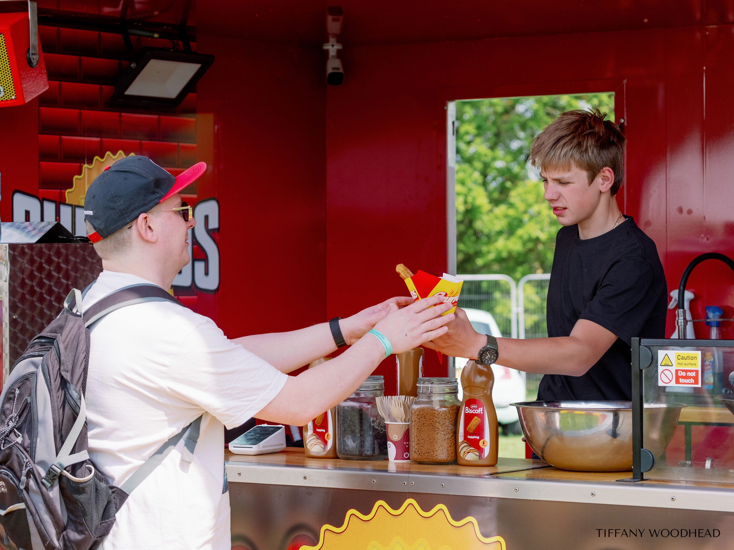 A young man with a backpack purchasing food from a vendor at a food stand or truck, with jars of toppings and condiments on the counter.